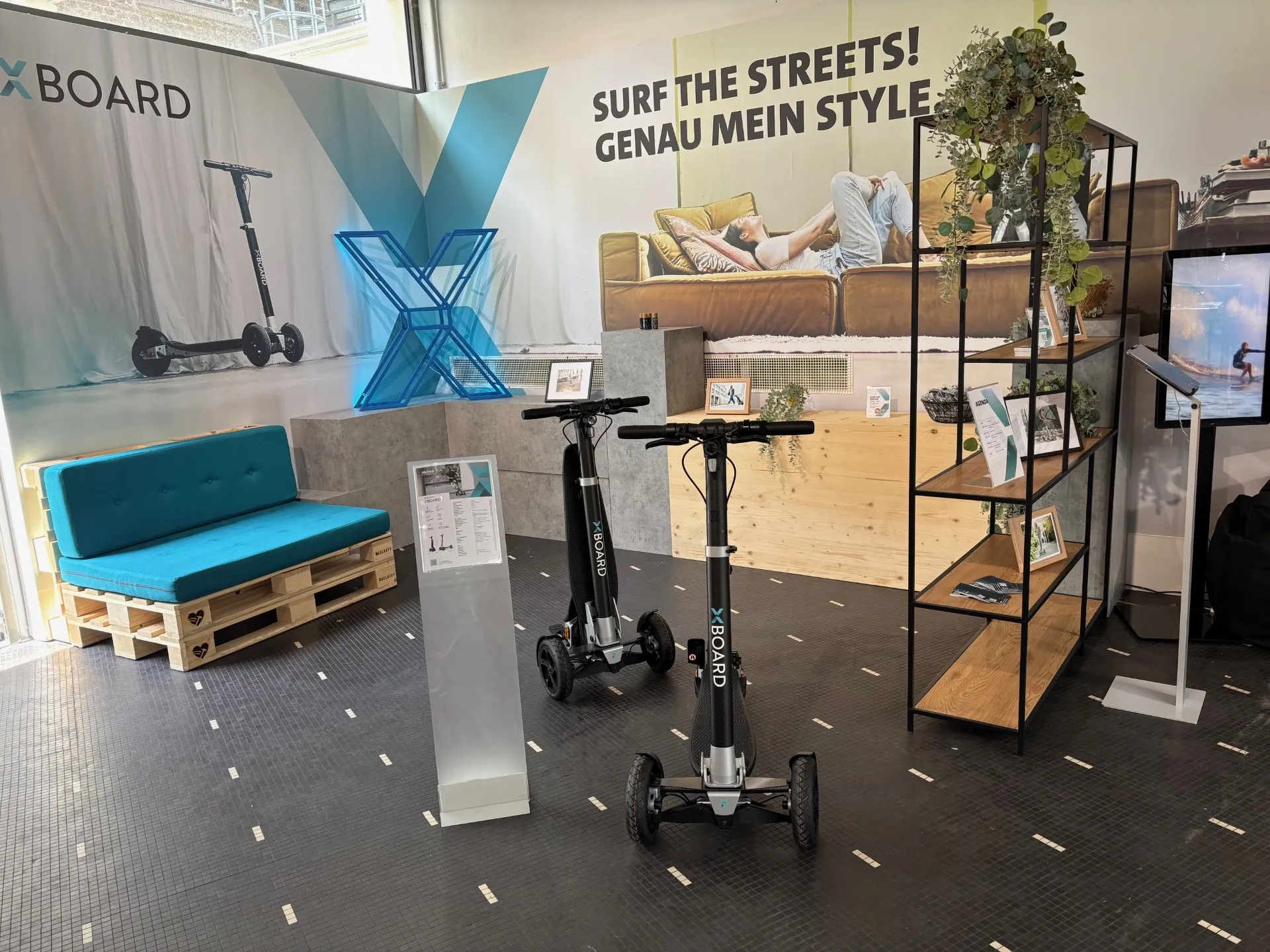 XBoard-Showroom mit zwei E-Scootern, modernem Lounge-Bereich, Regal und Surf-Motto im Hintergrund.