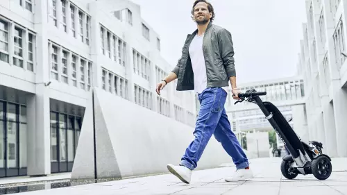 A man in casual clothing, wearing jeans, a t-shirt, and a jacket, walks through a modern urban environment while pulling an e-scooter behind him. In the background, tall white buildings with many windows are visible.