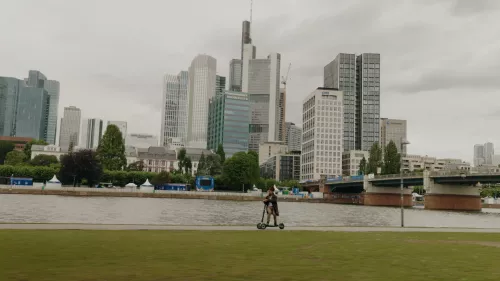 A person rides an XBOARD electric scooter along a riverside path, with a backdrop of modern skyscrapers and buildings. The scene includes a calm river, green grass, and an overcast sky. The city skyline and bridge are prominent in the background.
