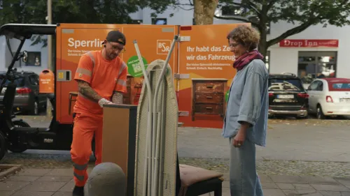 BSR-Mitarbeiter lädt Sperrling-Cargobike; Kundin übergibt Sperrmüll (Bügelbrett, Möbel)