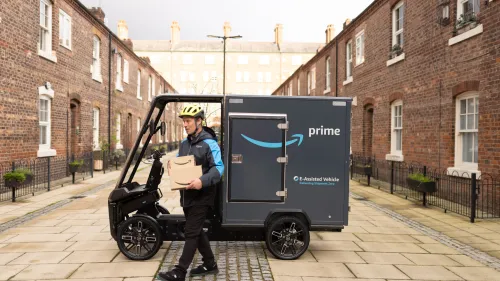 Amazon-Zusteller verlässt E-Cargobike mit Paket in gepflasterter Wohnstraße zwischen Backsteinhäusern.