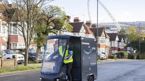 Amazon-E-Cargobike auf Straße in Wohngegend, Fahrerin mit Warnweste beim Zustellen von Paketen.