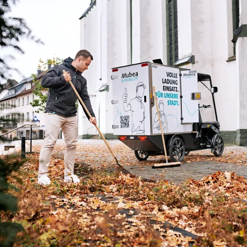 Ein junger Arbeiter kehrt herbstliches Laub vor einem E-Cargobike.