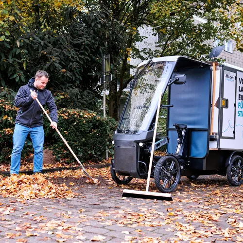 Young worker sweeping leaves, in the background is his eCargobike with other street cleaning tools.