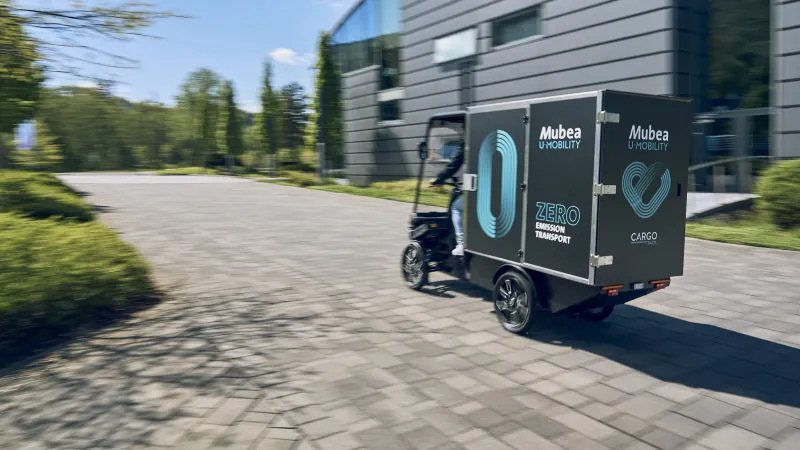 E-Cargobike mit großem Laderaum fährt auf gepflastertem Weg an modernen Gebäude entlang.