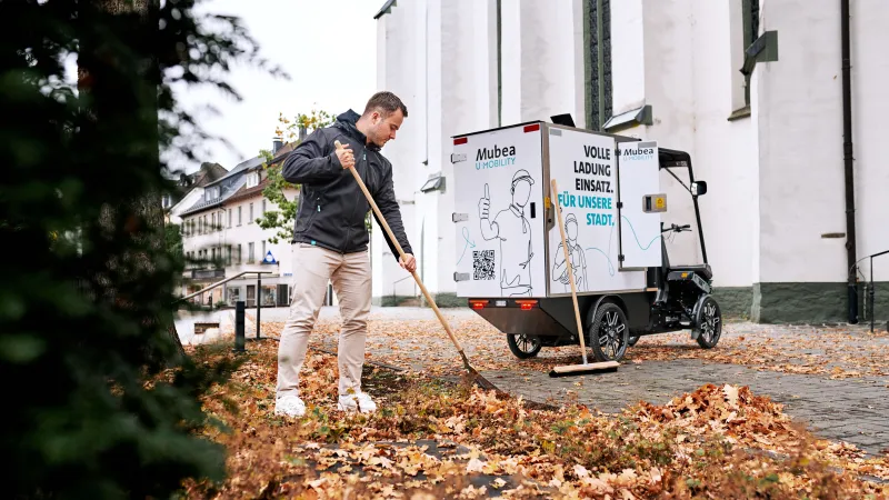 A young worker sweeps autumn leaves in front of an eCargobike.
